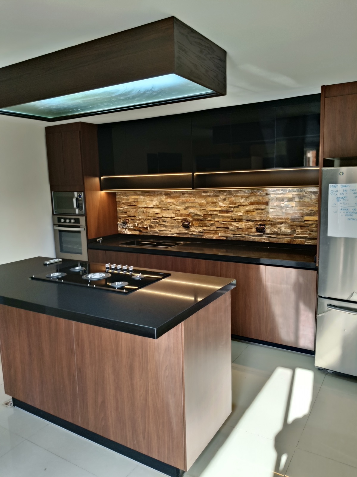 Cocina con isla de madera, cubierta negra, backsplash de piedra laja y plafón iluminado